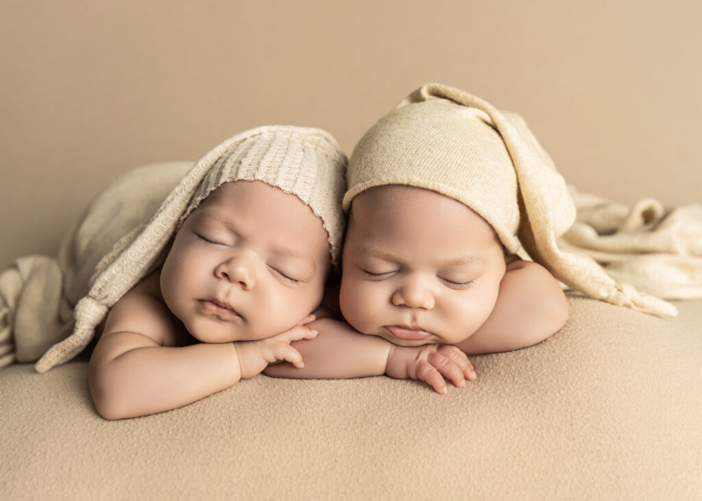 Twins snuggled together wearing cream hats during a newborn photo session.